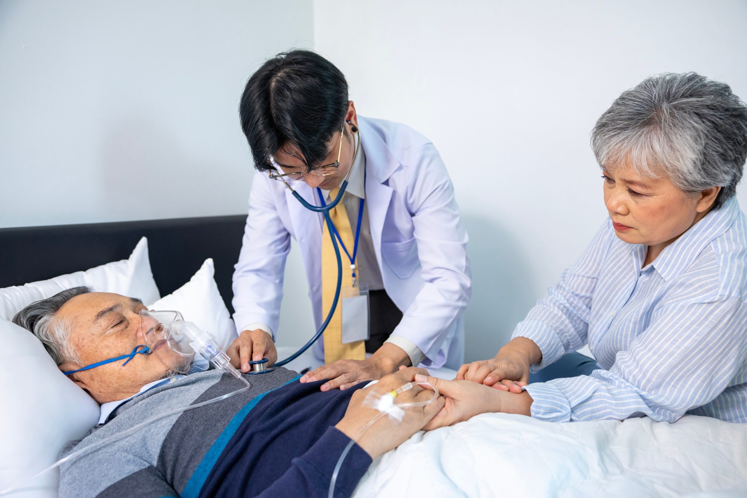 Paciente masculino de edad avanzada en cama siendo consolado por su esposa mientras es examinado por su joven médico 