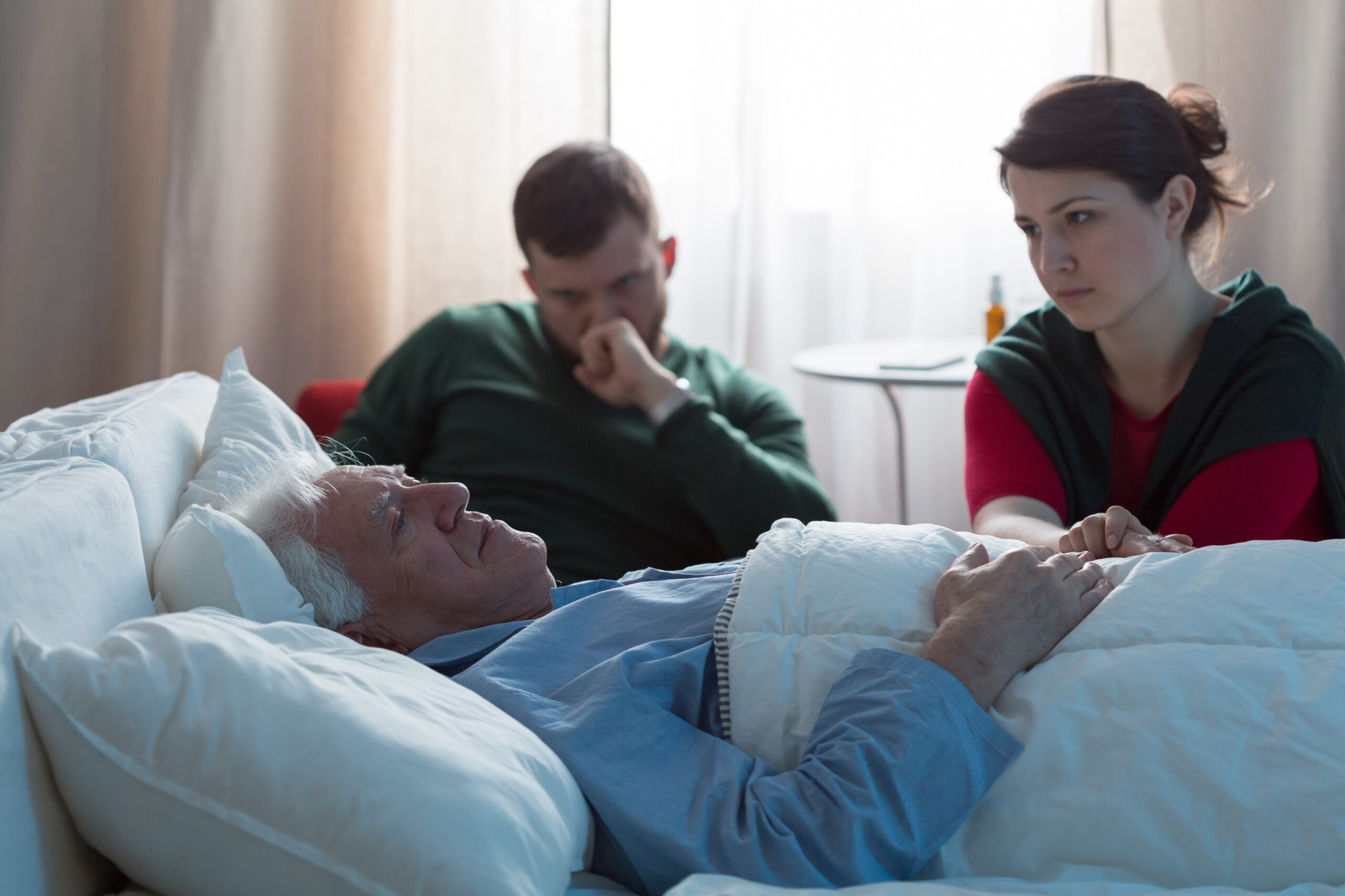 Paciente masculino de edad avanzada en la cama mientras su hijo y su hija están sentados cerca de él 